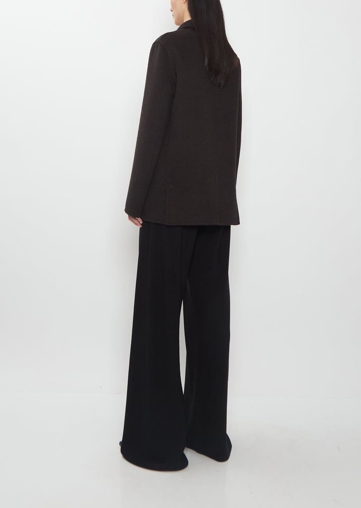 Rear view of the dark brown The Row Cinna Cashmere Jacket paired with black trousers.