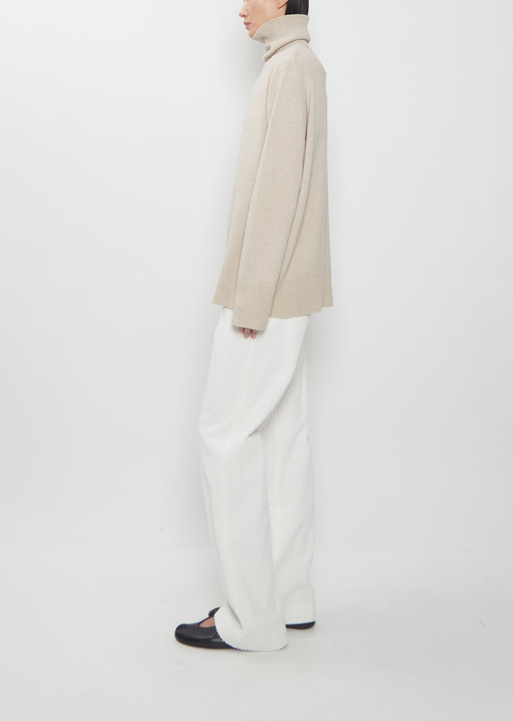 Side view of The Row Stepny Wool Cashmere Top in sand beige worn with white trousers.