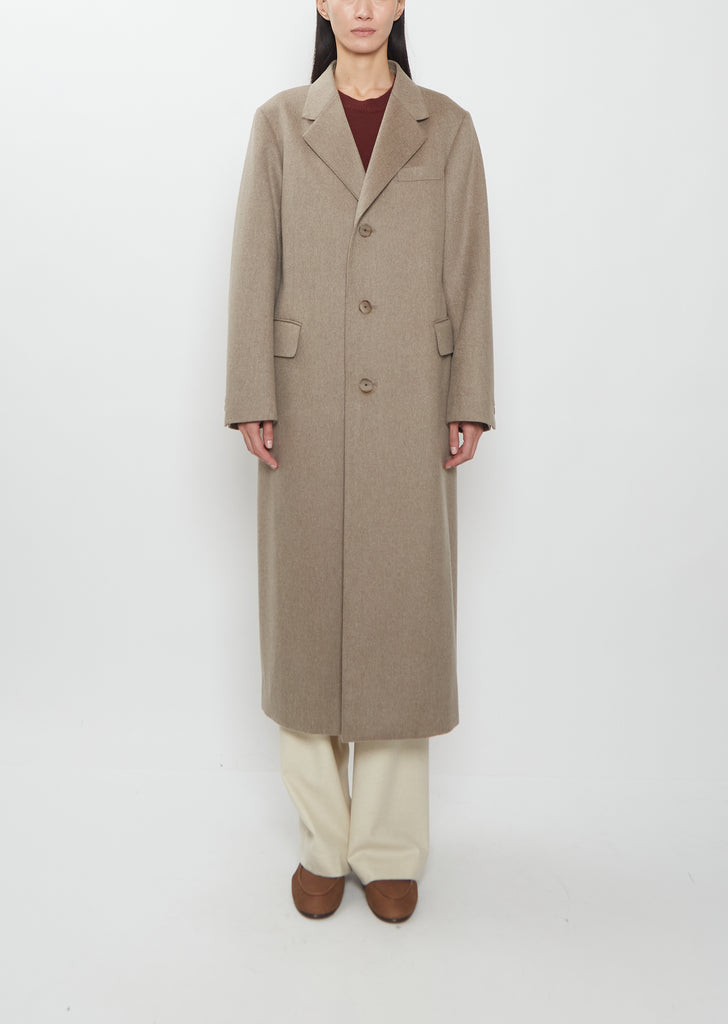 Front view of a model wearing the Auralee cashmere moleskin long coat in light brown over cream trousers.