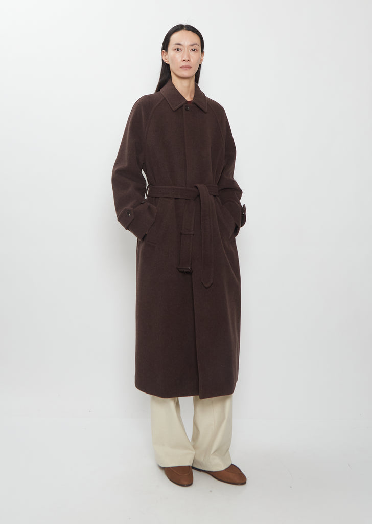 A person wearing a long, belted dark brown Super Fine Wool Mosser Soutien Collar Coat over light-colored trousers.