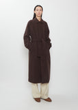 A person wearing a long, belted dark brown Super Fine Wool Mosser Soutien Collar Coat over light-colored trousers.