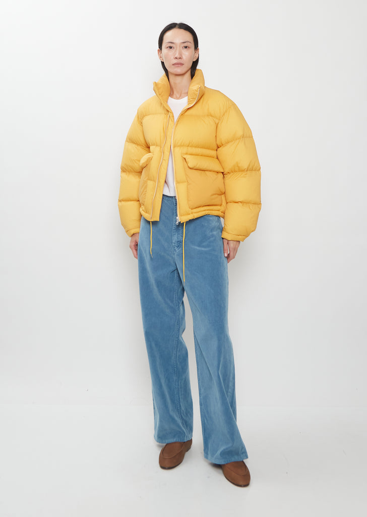 A person wears a yellow Super Light Nylon Ripstop Down Blouson, white t-shirt, blue trousers, and brown shoes.