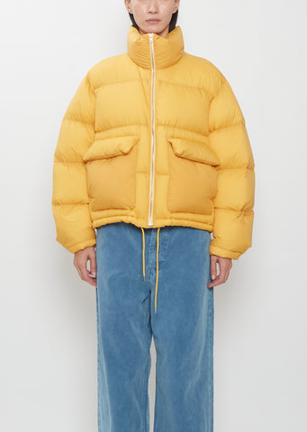 A person wearing a yellow Super Light Nylon Ripstop Down Blouson with two large front pockets and blue trousers.