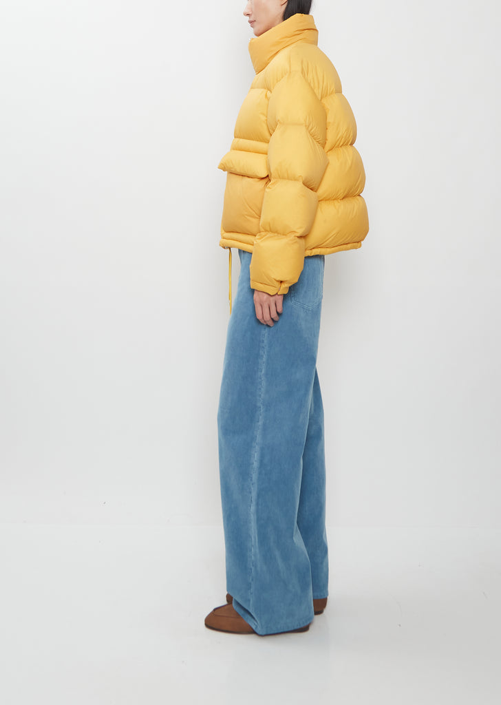 A person wearing a bright yellow, boxy Super Light Nylon Ripstop Down Blouson and loose-fitting blue trousers.